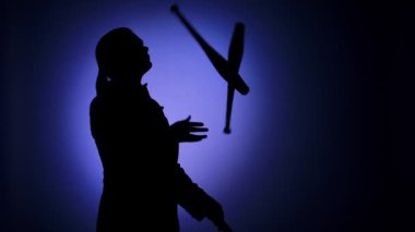Siyah siluet, spot ışıklarındaki iğnelerle hokkabazlık yapan bir adam. Eğlence programı olan bir sirk göstericisi. Stüdyo ışıklarıyla aydınlatılan mavi arka plan. Kapatın. Yavaş çekim.