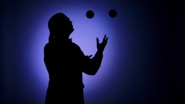 Siyah siluet, spot ışıklarında top oynayan bir adam. Eğlence programı olan profesyonel bir sirk göstericisi. Stüdyo ışıklarıyla aydınlatılan mavi arka plan. Kapatın. Yavaş çekim.