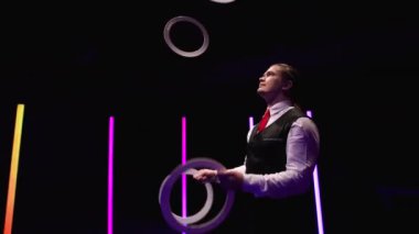 Kamera, beyaz halkalarla hokkabazlık yapan profesyonel bir erkek hokkabazın etrafında dönüyor. Tarz sahibi bir adam, parlak neon ışıkların arasında siyah stüdyo geçmişine karşı yeteneklerini ve tepkilerini bileyebilir. Kapatın. Yavaş çekim.