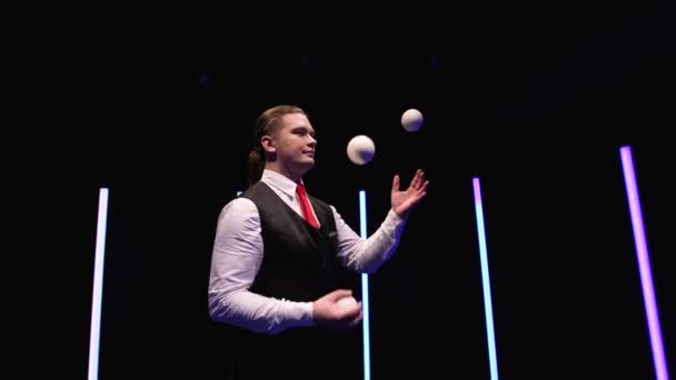Камера кругового руху навколо професійного циркового виконавця жонглює білі кульки на чорному студійному фоні серед яскравих неонових вогнів. Близько. Повільний рух . — стокове відео