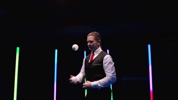 Cámara de movimiento circular alrededor de un artista de circo profesional haciendo malabares con bolas blancas contra un fondo de estudio negro en medio de luces de neón brillantes. De cerca. Movimiento lento. — Vídeo de stock