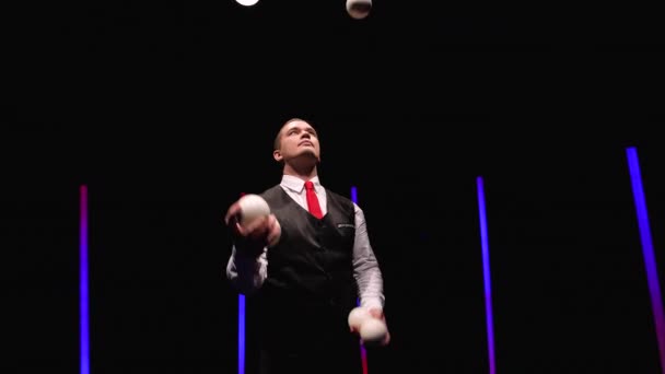 Artista de circo profesional de tiro orbital haciendo malabares con cinco bolas blancas contra un fondo de estudio negro en medio de luces de neón brillantes. De cerca. Movimiento lento. — Vídeos de Stock