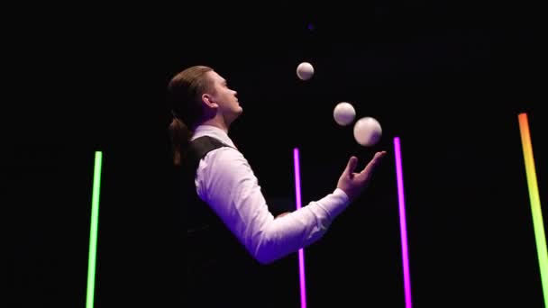 Orbitale tourné artiste de cirque professionnel jonglant avec cinq boules blanches sur un fond de studio noir au milieu de lumières au néon lumineux. Ferme là. Mouvement lent. — Video