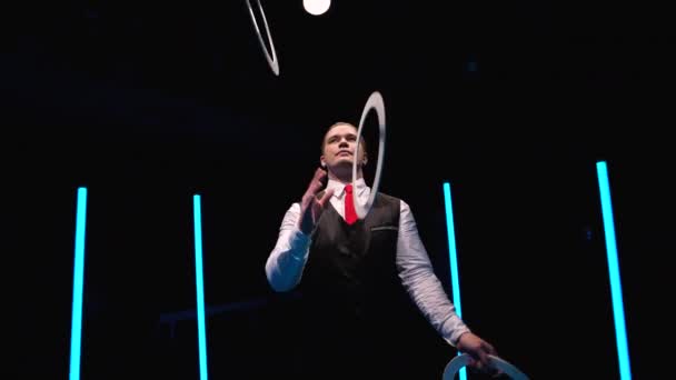 Camera rotates around professional male juggler juggling white rings. Stylish man hones his skill and reaction against black studio background amidst bright neon lights. Close up. Slow motion. — Stock Video