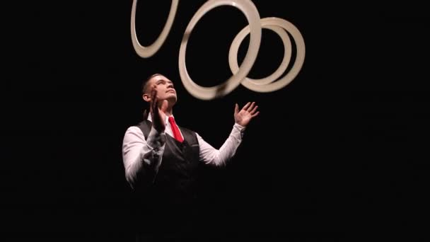 Camera rotates around concentrated man juggling white rings, then puts them around his neck. Juggler shows tricks on a black studio background. Orbital shot close up. Slow motion. — Stock Video
