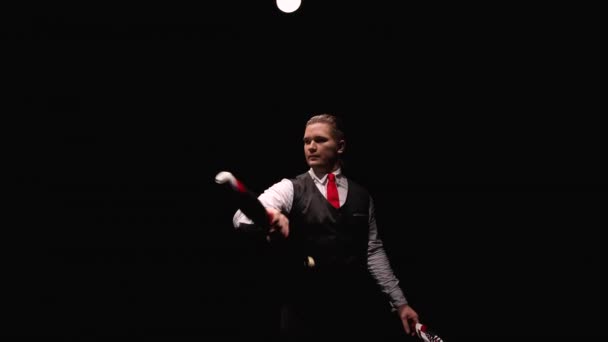 Kamera se otáčí kolem koncentrovaného muže profesionálně žonglujícího a balancujícího kolíky. Žonglér ukazuje triky na pozadí černého studia. Orbitální střela zblízka. Zpomalený pohyb. — Stock video