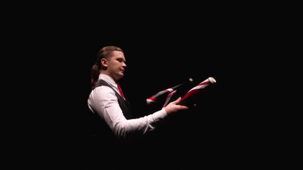 Orbital shot of a stylish man juggling with pins. The juggler hones his skill and dexterity against a black studio backdrop illuminated by lights. Professional circus show. Close up. Slow motion. — Stock Video