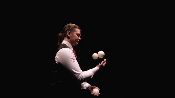 Orbital skott av en snygg man professionellt jonglera vita bollar. Cirkus artist upplyst av ljus kastar och fångar bollar på svart studio bakgrund. Närbild. Långsamma rörelser. — Stockvideo