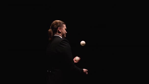 Orbitale Aufnahme eines Mannes im schwarzen Anzug, der professionell mit weißen Bällen jongliert. Ein Zirkuskünstler wirft und fängt, beleuchtet von Lichtern, in einem dunklen Studio Bälle. Aus nächster Nähe. Zeitlupe. — Stockvideo