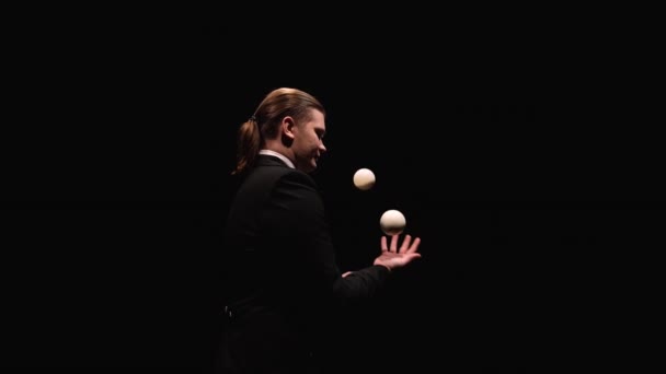 Orbital shot dari seorang pria dalam setelan hitam profesional juggling bola putih. Seorang pemain sirkus, diterangi oleh lampu, melempar dan menangkap bola di studio gelap. Tutup. Gerakan lambat. — Stok Video