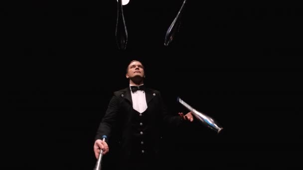 Camera rotates around circus juggler using pins to juggle. A man in a black suit performs exciting tricks in a dark backlit studio. Orbital shot close up. Slow motion. — Stock Video