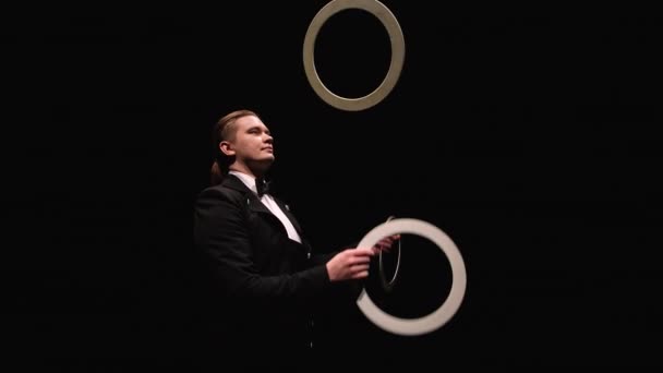 La fotocamera ruota intorno showman, spatola giocoleria anelli bianchi. Juggler, illuminato da luci, mostra trucchi su uno sfondo nero da studio. Colpo d'orbitale da vicino. Rallentatore. — Video Stock