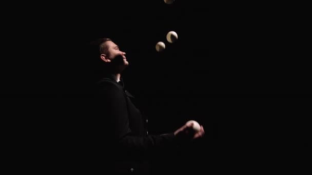 Orbitální záběr muže v černém obleku profesionálně žonglujícího s bílými koulemi. Cirkusový účinkující, osvětlený světly, hází a chytá míče v temném studiu. Zavřít. Zpomalený pohyb. — Stock video