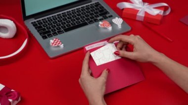 Kadın eli, sevgililer günü kartpostalını bir zarfa koyup dekoratif bir kalple süslemiş. Dizüstü bilgisayarı ve hediye kutuları olan kırmızı bir masa. Kapatın. Yavaş çekim.
