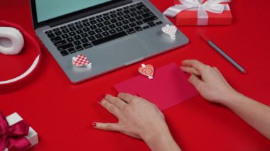 Kadınların elleri dekoratif bir kalple süslenmiş bir zarfı açar ve bir aşk kartı çıkarır. Dizüstü bilgisayarı ve hediye kutuları olan kırmızı bir masa. Sevgililer Günü. Kapatın. Yavaş çekim