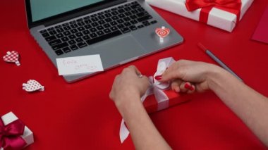 Kadınların elleri hediye paketini sarıp beyaz fiyonk bağlıyor. Dizüstü bilgisayarlı kırmızı masa, hediyeler, kalpler ve sevgililer günü kartı. Sevgililer bayramına hazırlanıyorum. Sevgililer Günü konsepti. Kapatın. Yavaş çekim