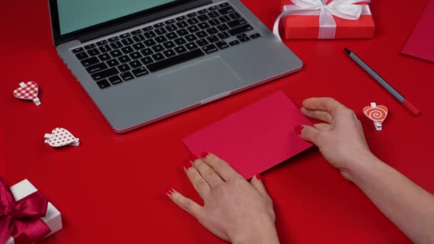 Le mani di donne aprono la busta festiva e tirano fuori la carta d'amore di San Valentino con l'iscrizione La amano. Vista dall'alto del tavolo rosso con computer portatile e scatole regalo. Il concetto di San Valentino. Chiudete. Rallentatore. — Video Stock