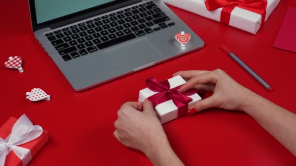 Le mani delle donne disfanno la confezione regalo, annullano l'arco. Vista dall'alto del tavolo rosso con computer portatile, regali, cuori e San Valentino. Umore romantico. Il concetto di San Valentino. Chiudete. Rallentatore. — Video Stock