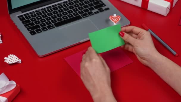 Les mains des femmes mettent dans l'enveloppe festive une carte postale d'amour de la Saint-Valentin avec une touche chromatique à écran vert. Vue du dessus de la table rouge avec un ordinateur portable et des boîtes-cadeaux. Concept de Saint-Valentin. Ferme là. Mouvement lent. — Video