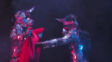 Actors in shiny costumes of bulls from mirrors playfully box and butt horns on a red rag. Bulls in reflective suits twinkle in colored lights against a black studio background. Close up. Slow motion.