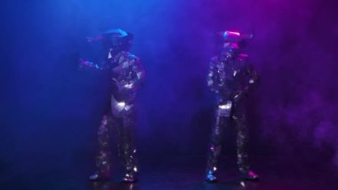 Parlak aynalı kostümlü, boğa kafalı ve boynuzlu iki aktör festival partisinde dans ediyor. Koyu renkli dumanlı stüdyodaki renkli pembe neon ışıkların arka planında yansıyan insanları göster. Yavaş çekim.