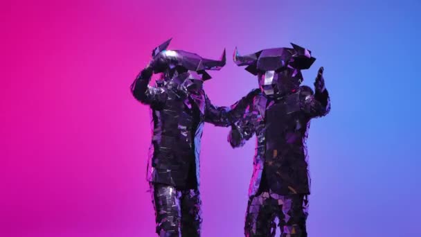 Two people in futuristic costumes of bulls from the mirrors cheerfully wave inviting them to come here to dance. Symbol of 2021, the bull, shines and is reflected in the light. Slow motion. Close up. — Stock Video