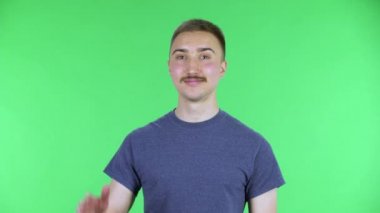 Kameraya bakıp el sallayan ve jest yapan genç bir adamın portresi. Bıyıklı, mavi tişörtlü, stüdyoda yeşil ekranda poz veren yakışıklı bir erkek. Kapat..