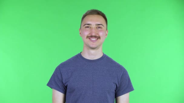 Egy fiatal férfi portréja, aki a kamerába néz és mosolyog. Aranyos férfi, bajusszal, kék pólóban, zöld vásznon pózol a stúdióban. Közelről.. — Stock videók