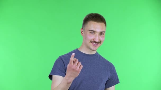 Ritratto di un giovane uomo che guarda la macchina fotografica salutando scherzosamente e facendo un gesto venire qui. Carino maschio con i baffi in una t-shirt blu in posa su uno schermo verde in studio. Da vicino.. — Video Stock