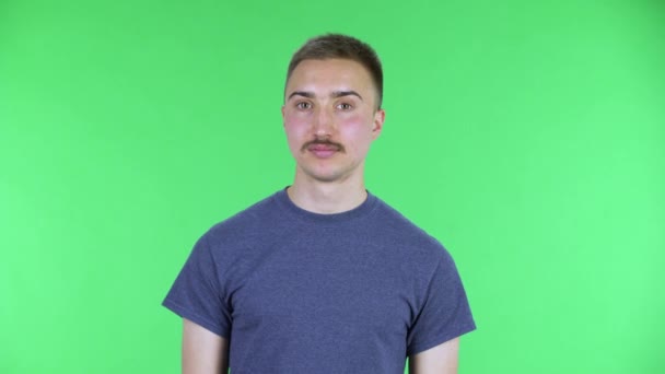 Portret van een jongeman die naar de camera kijkt zegt dat hij geschokt is. Leuke man met een snor in een blauw t-shirt poserend op een groen scherm in de studio. Sluiten.. — Stockvideo