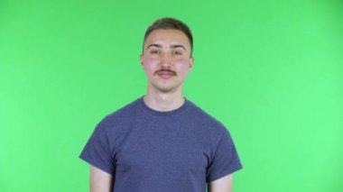 Kameraya bakan genç bir adamın portresi. Dost canlısı dalgalar. Buraya gelen jestler. Bıyıklı, mavi tişörtlü, stüdyoda yeşil ekranda poz veren yakışıklı bir erkek. Kapat..