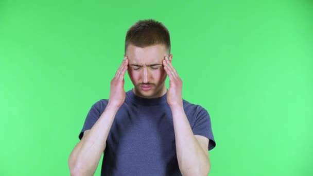 Portrait d'un jeune homme souffrant de maux de tête et se frotte les tempes. Mignon mâle avec une moustache dans un t-shirt bleu posant sur un écran vert dans le studio. Gros plan. — Video