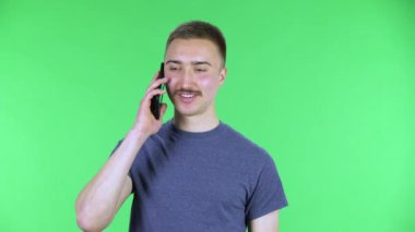 Cep telefonu için konuşan ve sevinen genç bir adamın portresi. Bıyıklı, mavi tişörtlü, stüdyoda yeşil ekranda poz veren yakışıklı bir erkek. Kapat..