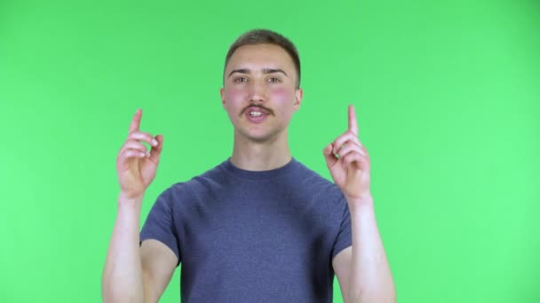 Porträtt av en ung man som pekar uppåt och skakar på huvudet. Söt man med mustasch i en blå t-shirt som poserar på en grön skärm i studion. Närbild. — Stockvideo