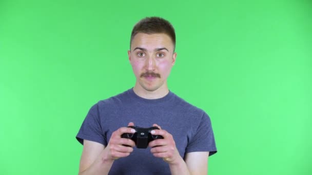 Portrait d'un jeune homme jouant à un jeu vidéo en utilisant une manette sans fil avec joie et se réjouissant de la victoire. Mignon mâle avec une moustache dans un t-shirt bleu posant sur un écran vert dans le studio. Gros plan — Video