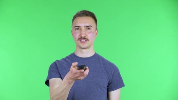 Retrato de un joven con TV a distancia en la mano, encendiendo la televisión y muy riendo, asustado y luego molesto. Lindo hombre con un bigote en una camiseta azul posando en una pantalla verde en el estudio. De cerca. — Vídeo de stock