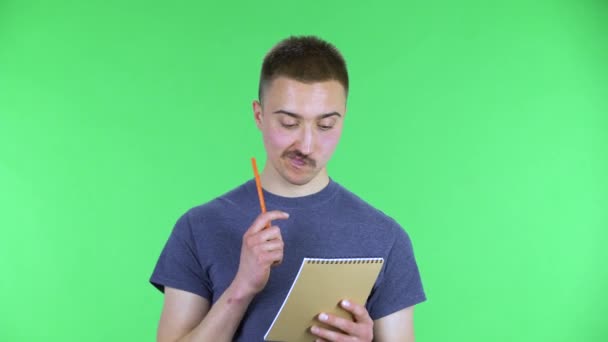 Portrait d'un jeune homme réfléchit, puis heureux d'écrire avec un crayon dans un cahier. Mignon mâle avec une moustache dans un t-shirt bleu posant sur un écran vert dans le studio. Gros plan. — Video
