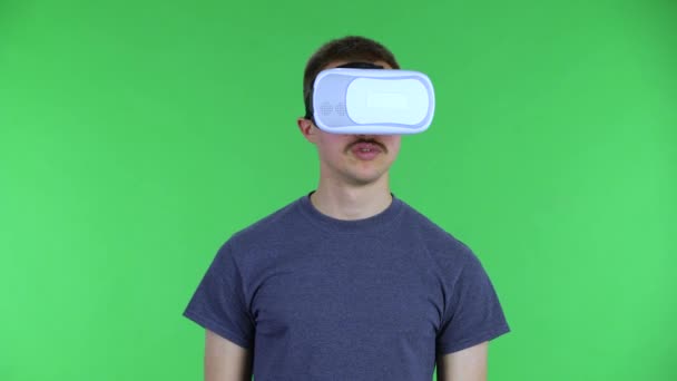 Retrato de un joven con auriculares de realidad virtual o gafas 3D. Lindo hombre con un bigote en una camiseta azul posando en una pantalla verde en el estudio. De cerca.. — Vídeos de Stock