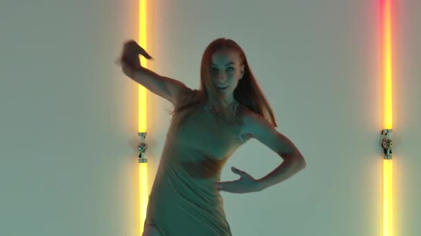 Mladá a energická žena v béžových dlouhých šatech tančí smyslné moderní opovržení. Štíhlá tanečnice se elegantně pohybuje v tmavém studiu na pozadí barevných neonových světel. Zavřít. Zpomalený pohyb. — Stock video