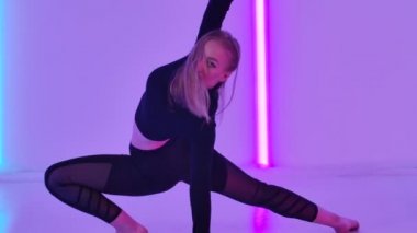 İnce dansçı tarafından icra edilen aerobik elementlerle, parlak neon ışıkların arka planına karşı duygulu ve modern bir aşağılama. Kadın, karanlık stüdyoda modern koreografi uyguluyor. Kapatın. Yavaş çekim.