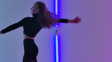 İnce dansçı tarafından icra edilen aerobik elementlerle, parlak neon ışıkların arka planına karşı duygulu ve modern bir aşağılama. Kadın, karanlık stüdyoda modern koreografi uyguluyor. Kapatın. Yavaş çekim.