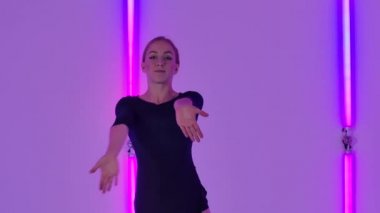 Tatlı, genç bir kadın zarif bir şekilde neon ışıkların arka planına karşı şehvetli bir küçümsemeyle zıplar ve döner. Siyah elbiseli ince dansçı, esnekliğini yavaş çekimde gösteriyor. Kapat..