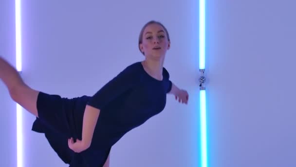 Lieve jonge vrouw springt en wervelt gracieus in sensuele minachting tegen een achtergrond van heldere neon lichten. Slanke danseres in zwarte jurk demonstreert haar flexibiliteit in slow motion. Sluiten.. — Stockvideo