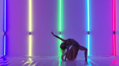 Bej uzun elbiseli modern bayan dansçı karanlık stüdyoda güzel plastik dramatik danslar yapıyor. Esnek dans eden kadın parlak renkli neon ışıkların arka planına karşı. Yavaş çekim.