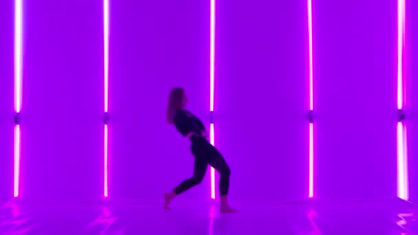 Flexibele danser voert acrobatische elementen van de moderne choreografie uit en springt tegen een achtergrond van paarse neonlichten. Sensuele vrouw oefent minachting uit in de schemerstudio. Langzame beweging. — Stockvideo