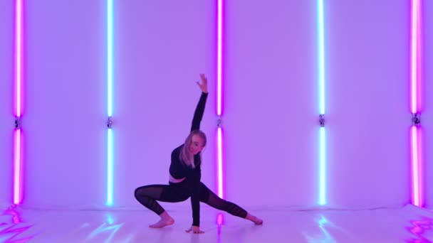 İnce dansçı tarafından icra edilen aerobik elementlerle, parlak neon ışıkların arka planına karşı duygulu ve modern bir aşağılama. Kadın, karanlık stüdyoda modern koreografi uyguluyor. Yavaş çekim. — Stok video