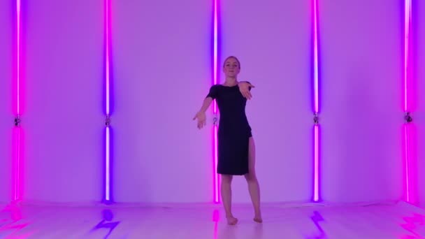 Dolce giovane donna salta e gira con grazia nel disprezzo sensuale su uno sfondo di luci al neon luminose. Ballerina snella in abito nero dimostra la sua flessibilità al rallentatore. — Video Stock
