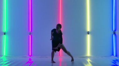 Siyah elbiseli ince bir dansçı tarafından yapılan akrobatik öğelere nazik bir aşağılama. Modern koreografiden hoşlanan genç ve güzel bir kadın karanlık stüdyoda parlak neon ışıkların önünde. Yavaş çekim.