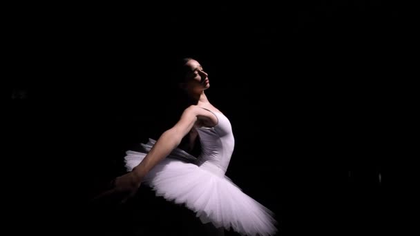 Camera rotates around gentle ballerina in a white tutu performing graceful movements with her hands. Orbital shot of a young dancer soaring against a black studio background. Close up. Slow motion. — стоковое видео