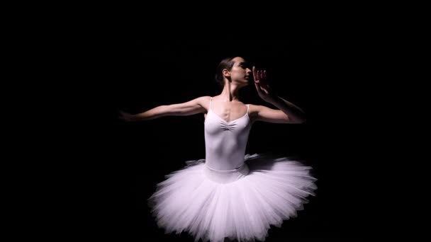 Camera rotates around gentle ballerina in a white tutu performing graceful movements with her hands. Orbital shot of a young dancer soaring against a black studio background. Close up. Slow motion. — стоковое видео